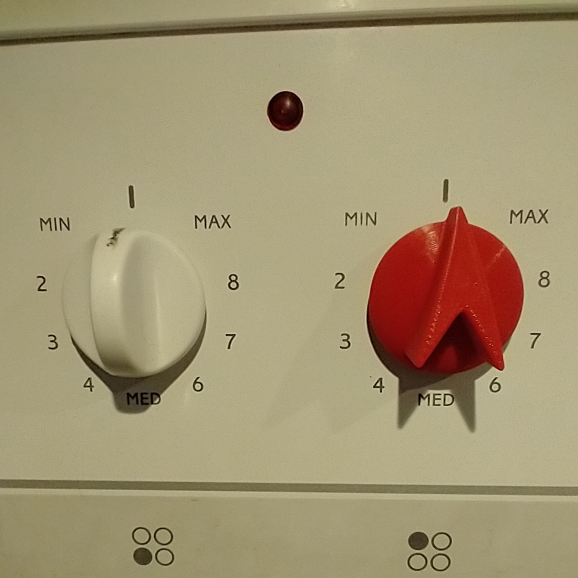 The panel of a stove with two knobs. On the left, a white knob has a vertical tab. There is a ragged black indicator line drawn on the top of the tab.. On the left, the red tab has an arrowhead profile with a clear indication of position.